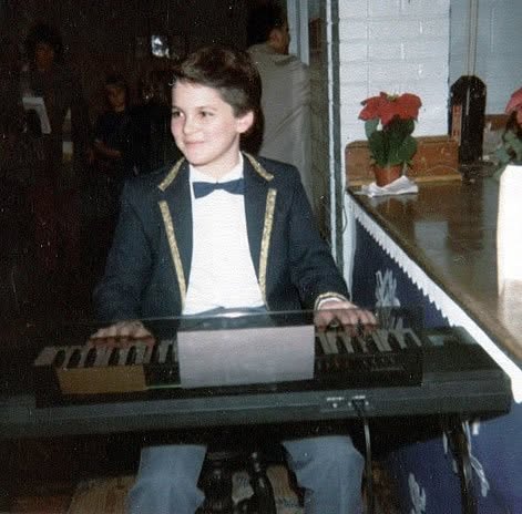 A smiling boy in a dark blazer with gold trim and a bow tie, seated at a small electronic keyboard with sheet music propped in front of him