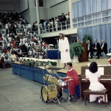 A high school graduation in an arena: a young Andrew in red cap and gown plays a gold drum kit on stage while another graduate accompanies on an upright piano and a robed singer performs at a microphone