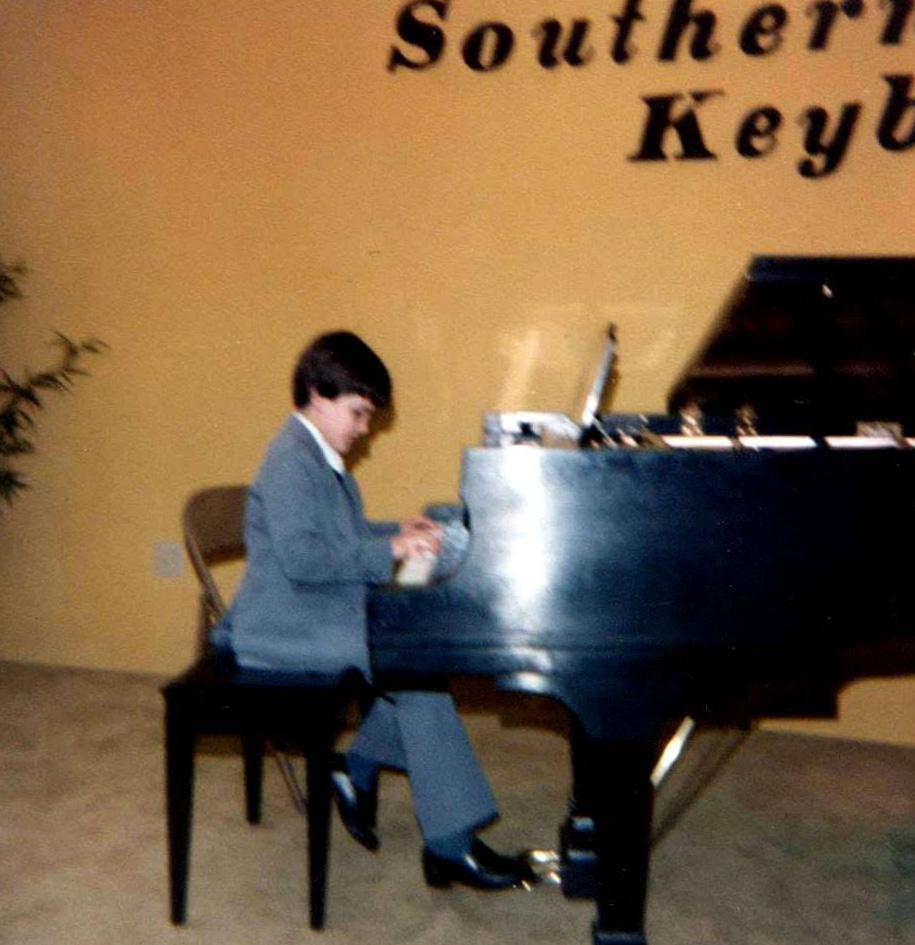 A young Andrew Doss in a gray suit, seated at a black grand piano and playing, with a Southern Keyboards sign on the gold wall behind him