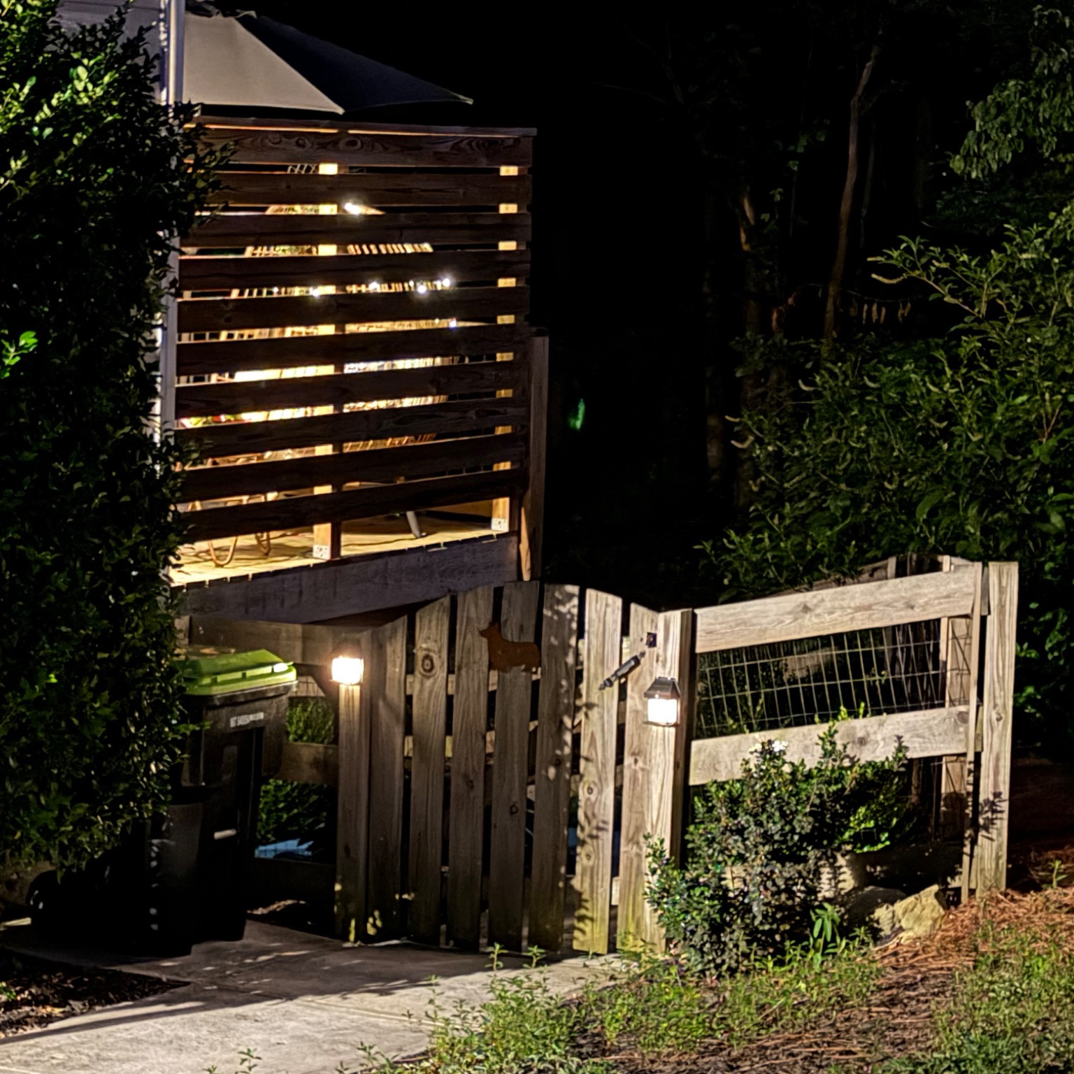 Warm light glowing through the slats of a wooden wall enclosing a deck at night, with small path lamps below and dark trees around