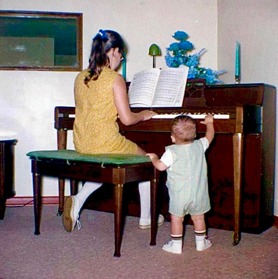 Andrew Doss as a toddler reaching up to touch the piano keys while his older sister Lisa plays
